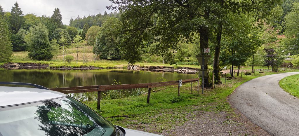 A lake with a road next to it. By the road, there's a gravel area, where a car is parked.