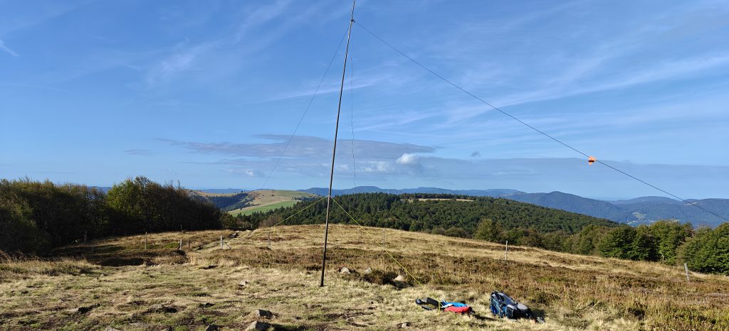 A grassy hilltop with some trees. Other hills are in the distance. My radio aerial mast and backpack are in the foreground.