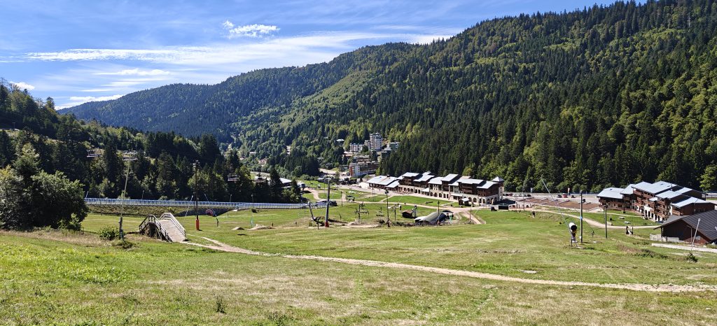 A green valley between forested hills. There are many buildings in the valley. The hill in the foreground has the apparatus of a ski resort, including chair lifts and snow blowers.